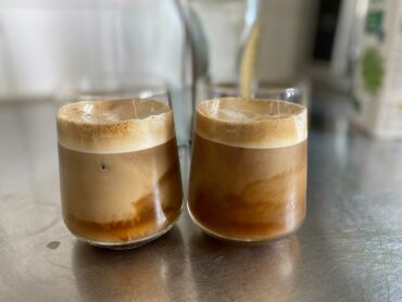 Två dricksglas står på en bänk och de är fyllda nästan till bredden med grekisk frappé som består av kaffe, is och mjölk