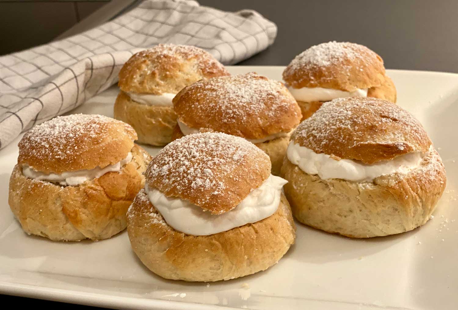 Ett stort vitt fat med 6 stycken veganska semlor står på en grå köksbänk. Bredvid fatet ligger en kökshandduk.