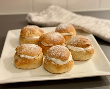 Ett stort vitt fat med 6 stycken veganska semlor står på en grå köksbänk. Bredvid fatet ligger en kökshandduk.