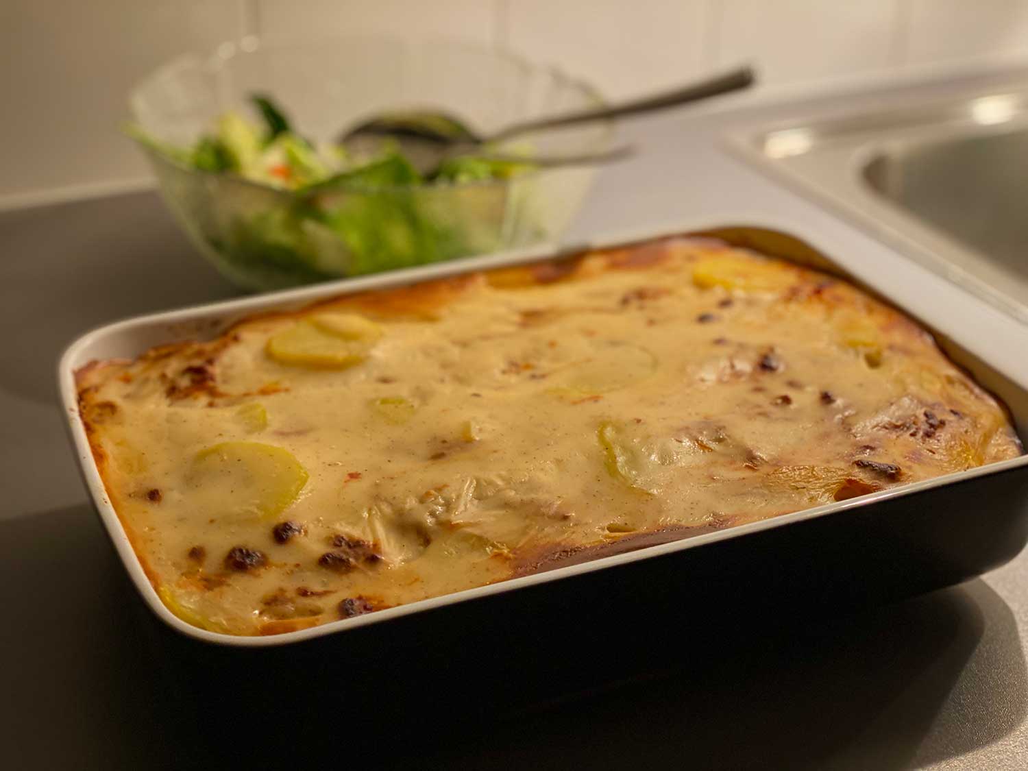 En ugnsform med vegansk moussaka med potatis står på en köksbänk. Bredvid formen står en salladsskål.