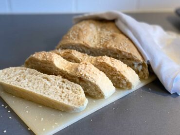 På en liten vit skärbräda ligger en limpa med snabbt lantbröd där tre brödbitar skurits upp så att man kan se insidan av brödet. Brödet har toppats med sesamfrön.
