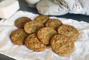 På köksbänken ligger en bakduk och på duken ligger massor av veganska Chocolate Chip Cookies. I bakgrunden skymtas en bunke med socker och en ungsvante.